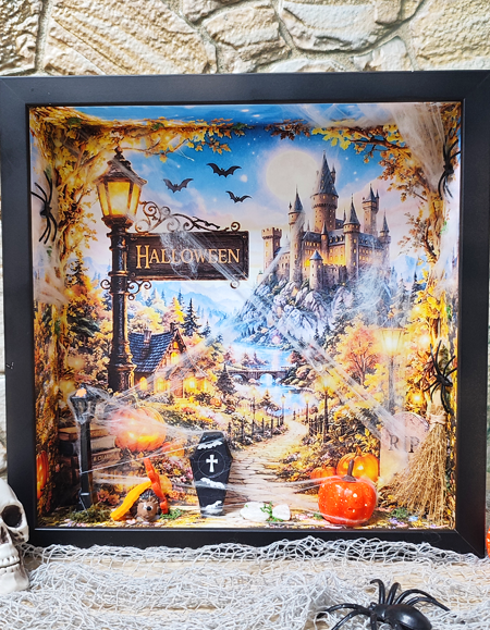 Kézműves őszi 3D árnyékdoboz: gótikus kastély és Halloween tábla, előtérben átszőtt fehér pókhálóval, narancssárga tökökkel és sírkővel.